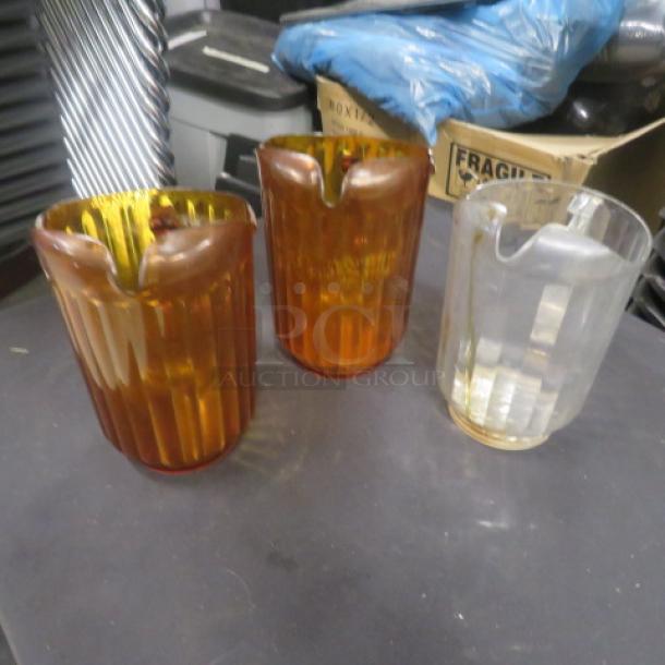 Assorted glass pitchers, two amber-colored and one clear, ribbed design, good condition, suitable for home decor or collection.