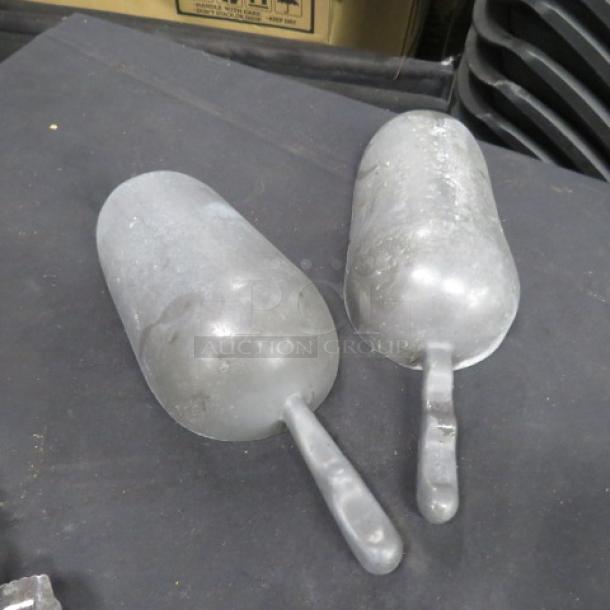 Two Aluminum Scoops with handles on dark surface; show signs of wear. Ideal for grain, feed, or ice.