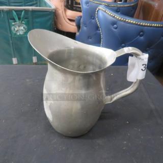 Stainless steel pitcher with a curved handle, showing signs of wear. Auction lot tag labeled "37" attached.