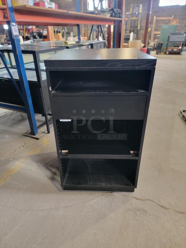 Matte black retail register stand with recessed shelf, smooth-glide drawer, and adjustable shelving. Durable for high-traffic use.