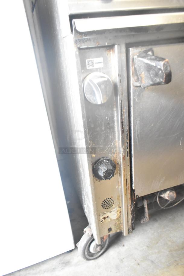 Commercial gas-powered 6 burner range with 2 tiers, oven, over shelf, and back splash. Features commercial casters. Visible wear.