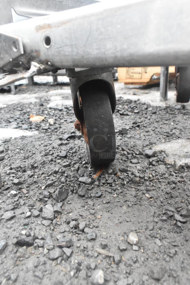 Two commercial pan transport racks on casters, outdoor setting, showing close-up of a wheel, indicating mobility and condition.