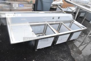 Brand new Regency 600S318181L 16 gauge stainless steel three-compartment sink with left drainboard, cross bracing, and tags indicating model details; minor scratches visible.