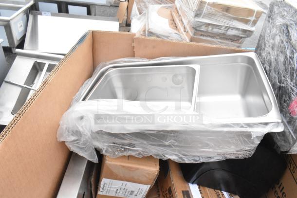 Box of 15 new scratch and dent items, including a stainless steel sink and splash guard, wrapped in plastic.