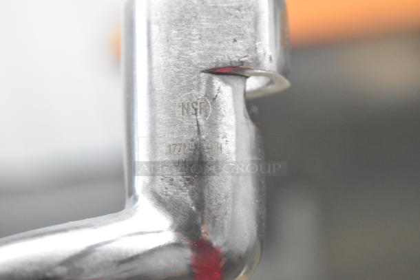Close-up of a metal attachment with visible "NSF" marking and model number. Part of a set including whisk, paddle, dough hook, blades.