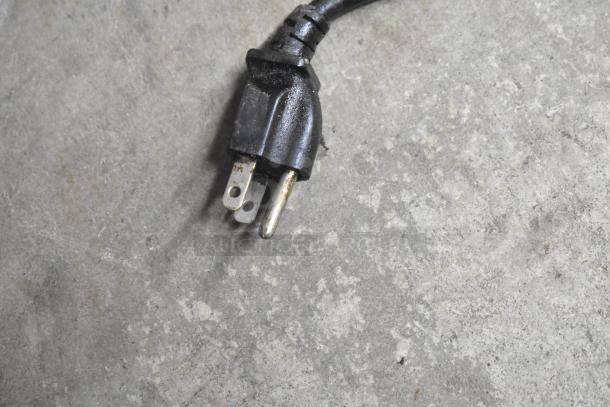 Close-up of a black three-prong power plug on a gray surface, part of the Hatco Heated Holding Merchandiser.