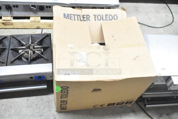 The image shows a cardboard box labeled "Mettler Toledo" on a concrete floor. The box is worn and partially open, revealing a metal object inside.