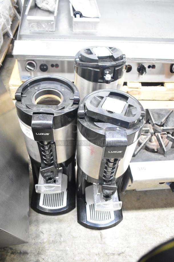 Three Fetco Luxus beverage dispensers on a countertop; stainless steel with black lids, labeled "Luxus," in good condition.