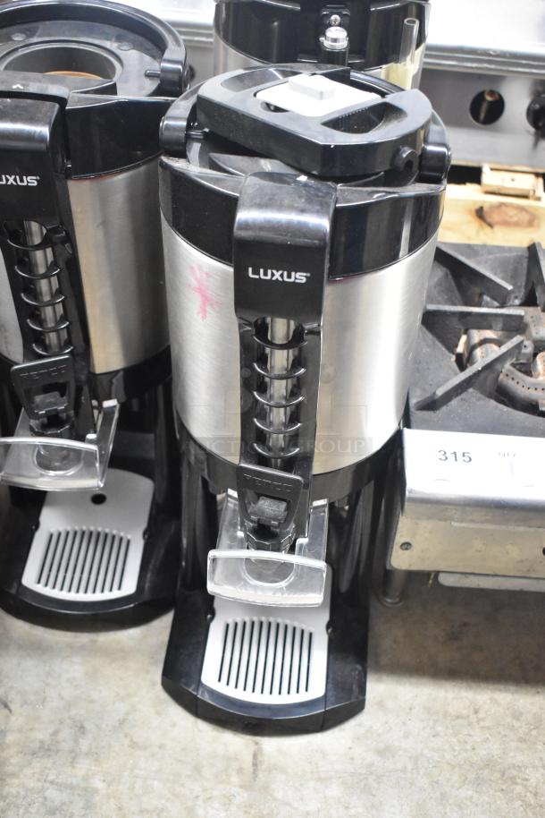 Three Fetco Luxus countertop beverage dispensers in stainless steel, each with a black handle and drip tray. Small pink mark visible.