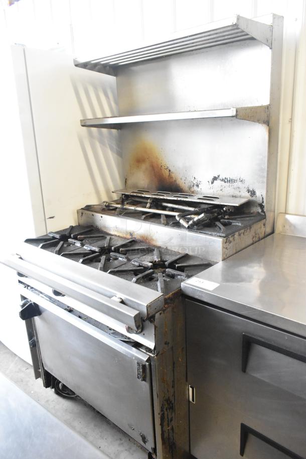 Commercial gas-powered 6 burner range with oven, over shelf, and backsplash on casters. Visible wear and rust on surfaces.