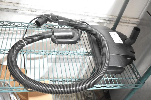 Vacuum cleaner attachment on metal shelf, features flexible hose and motor unit. Visible label reads "64". Condition appears used.