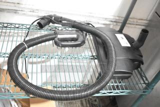 Vacuum cleaner attachment on metal shelf, features flexible hose and motor unit. Visible label reads "64". Condition appears used.