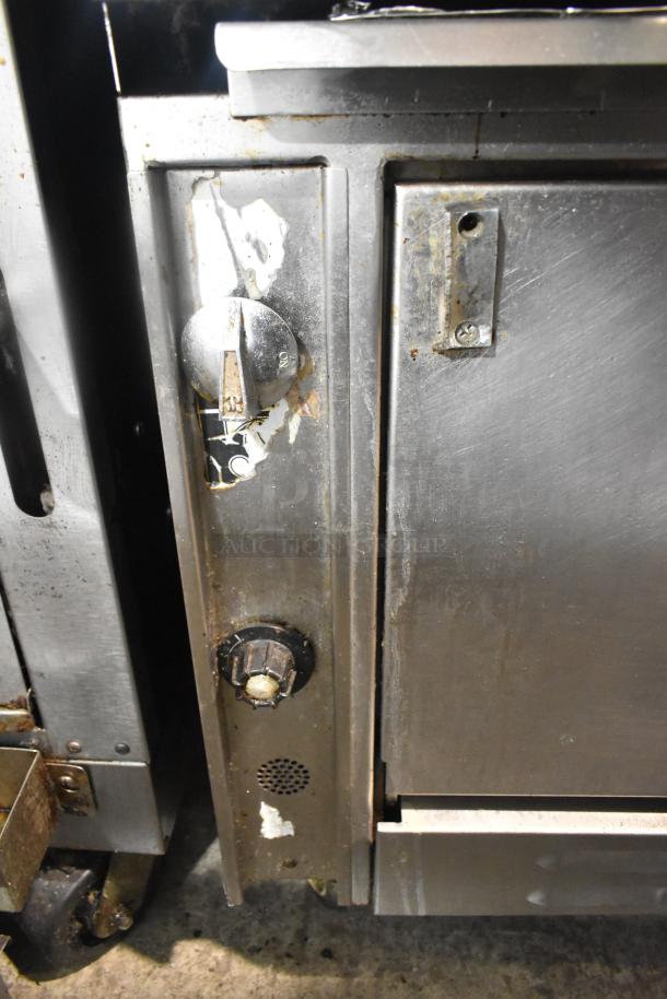 Commercial 12 burner gas range with 2 ovens, overhead shelf, backsplash, and casters. Visible wear, knobs intact.