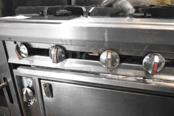 Commercial 12-burner gas range with two ovens, over shelf, back splash, and commercial casters; visible wear on knobs.