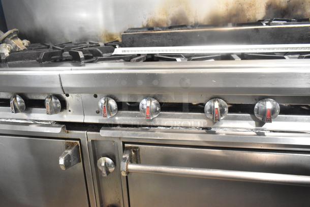 Commercial 12 burner gas range with dual ovens, over shelf, and backsplash on casters. Visible wear on knobs and surfaces.