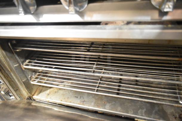 Commercial 6-burner range with salamander cheese melter and oven; stainless steel racks visible, used condition.