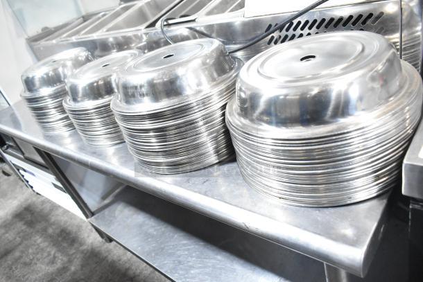 Stacks of metal dome covers on a stainless steel table. 98 pieces, shiny finish, used for food service.
