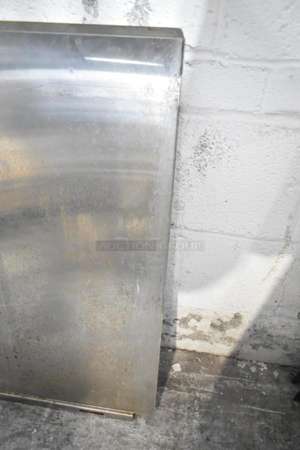 Alt text: Metal panel from auction lot, showing surface wear and slight rust, leaning against a white brick wall.