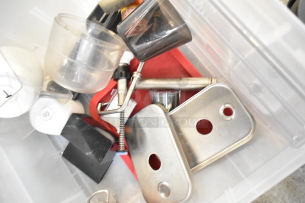 Various items in a clear plastic bin including metal brackets, a beverage dispenser part, and plastic bins. Mixed condition.