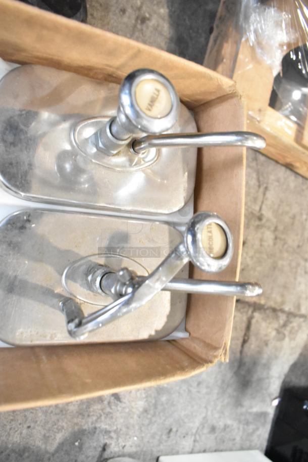 Two topping dispensers with labeled knobs "Vanilla" and "Chocolate" in a cardboard box, showing some wear.