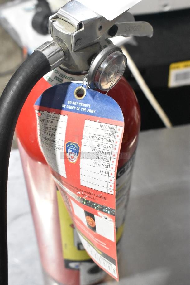 Metal fire extinguisher with attached inspection tag, serviced October 2023, requires buyer pickup, FDNY label visible.