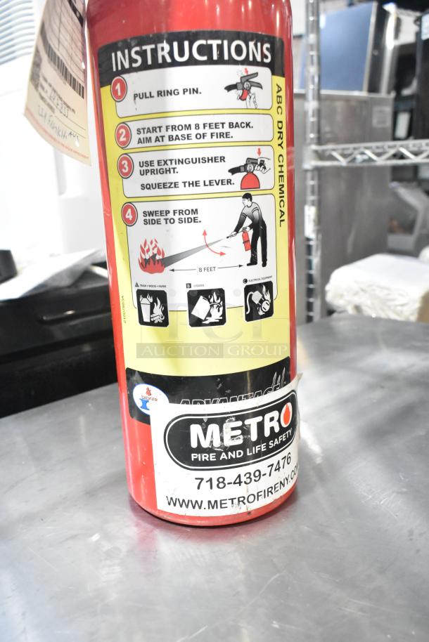 Red fire extinguisher with ABC dry chemical instructions. Label: Metro Fire and Life Safety, contact details visible.