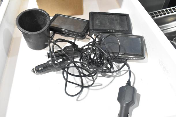 Four items including three Garmin GPS devices and one TomTom GPS, with tangled charging cables, in used condition.