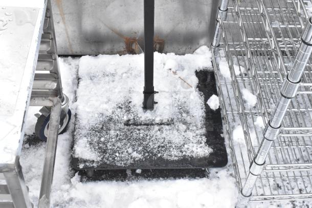 Outdoor sign base with a metal pole on a snowy surface, surrounded by metal shelving. Base is sturdy and weathered.