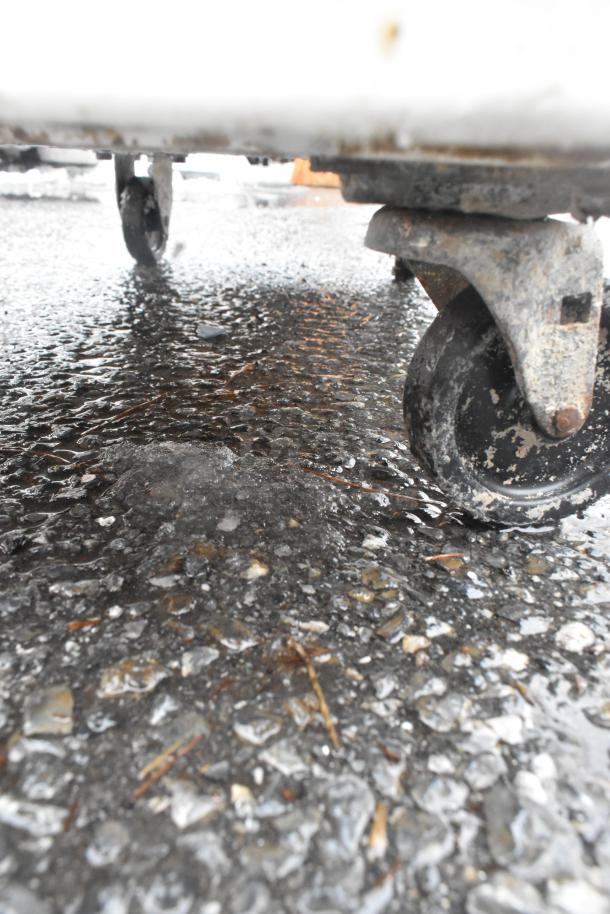 White poly ingredient bin on rusted commercial casters, outdoor setting, close-up of wheels, showing wear and usability.