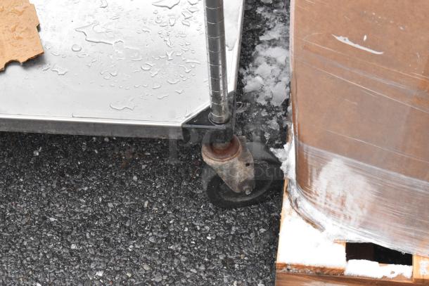 Stainless steel table with an under shelf on commercial casters, wet surface, showing part of the wheel and corner. Comes with tray stands.