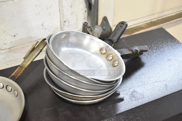 Stack of 11 metal skillets with riveted handles, showing signs of wear. Ideal for bulk purchase or cooking enthusiasts.