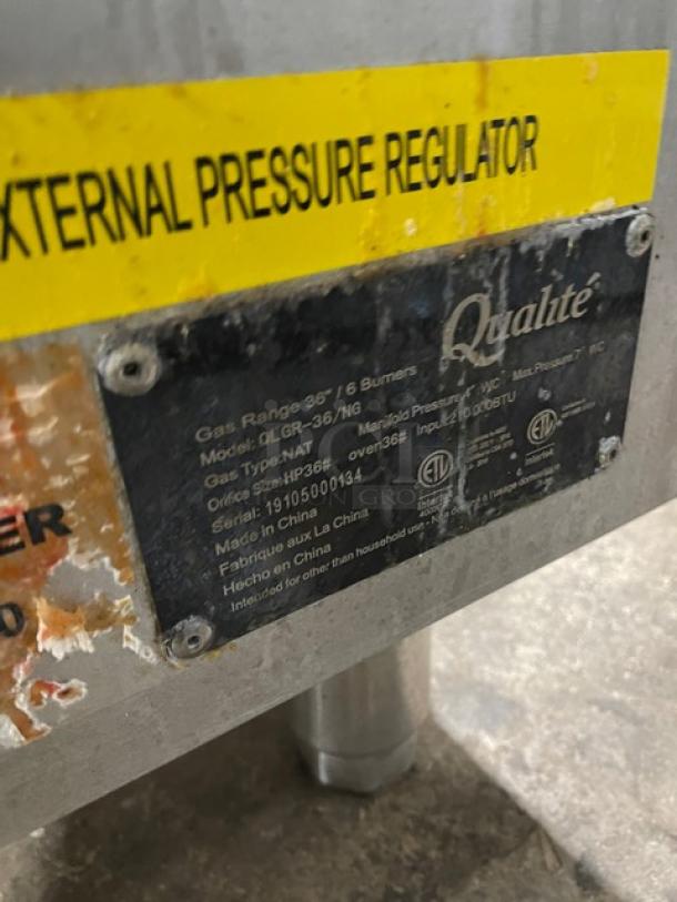 Qualite Commercial Stainless Steel Natural Gas 6-Burner Range/Oven, Model QLGR36NG, SN 19105000134. Features a backsplash, overhead salamander shelf, and wired oven rack. On legs. Labels detail model and serial numbers.