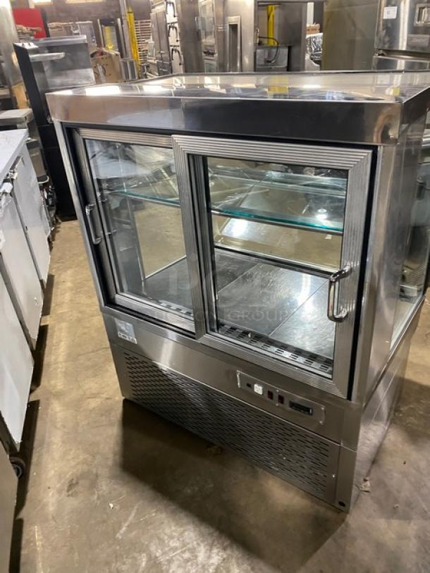 Commercial stainless steel refrigerated bakery display case with slanted glass, multi-tier shelving, and rear sliding glass doors.