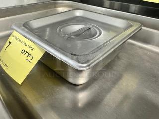 Lot of 2 stainless steel food pans with lids, slight wear, stacked on metal surface with marked auction tags.
