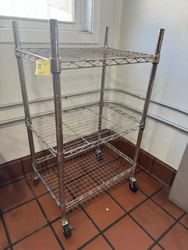 Chrome utility cart with three wire mesh shelves, caster wheels, and visible tag numbered 33, showing light wear.