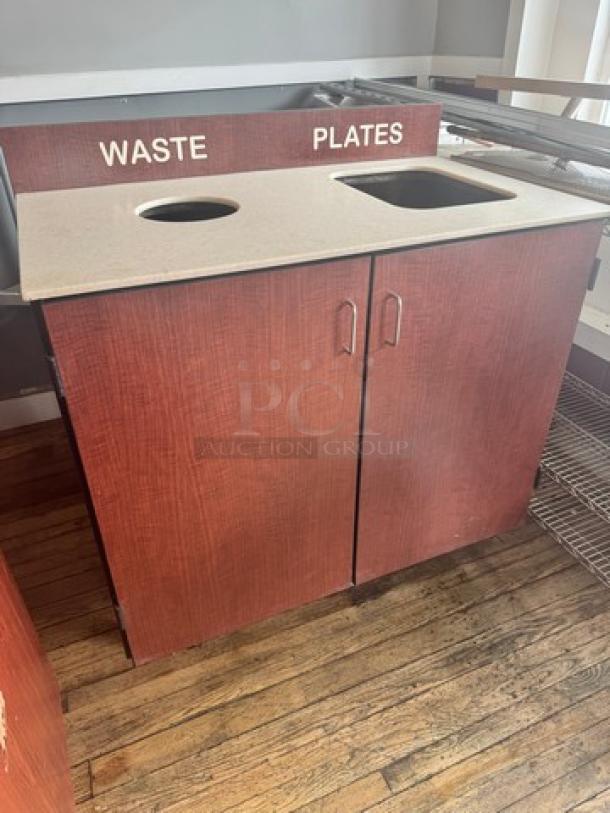 Wooden waste receptacle with dual openings labeled "WASTE" and "PLATES," features a cream countertop, in used condition.