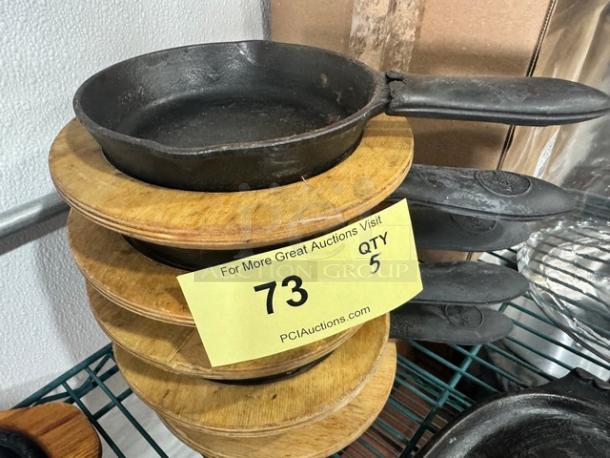 5" cast iron skillet BIDX5 with handle, stacked with wooden trivets, in good condition, auction label indicates quantity 5.