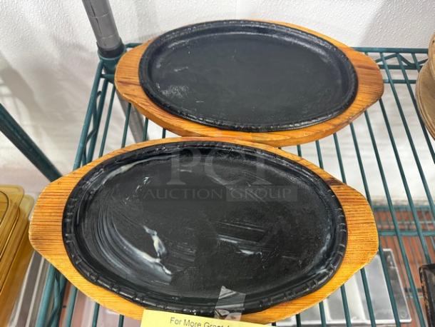 Pair of oval cast iron skillets with wooden bases, BIDX2 model, in used condition, suitable for serving sizzling dishes.