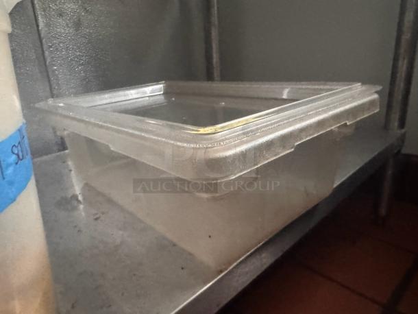 Clear plastic food storage container with lid, in used condition, on a metal shelf.
