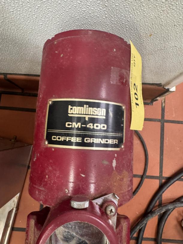 Tomlinson CM-400 commercial coffee grinder, maroon, visible wear. Label indicates model and brand. Auction tag attached.