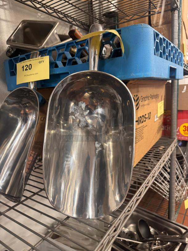 Stainless steel ice scoop on metal shelf, reflective surface, minor wear visible, number tag "120" attached.