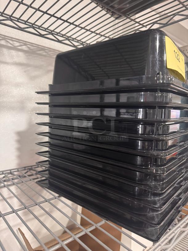 Stack of black plastic food pans BIDX10 on metal shelving. Each pan is clean and in good condition. Yellow label visible.