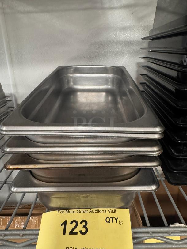 Stainless steel food pans, model BIDX6, in used condition, stacked on a shelf. Quantity of 6. Auction tag visible.