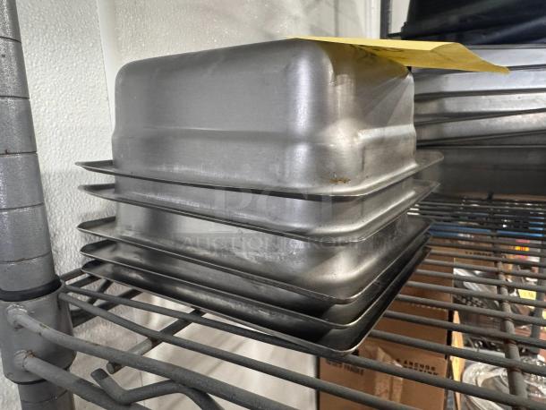 Stainless steel food pans BIDX5 stacked on metal shelves, showing some wear. Yellow paper label on top pan.