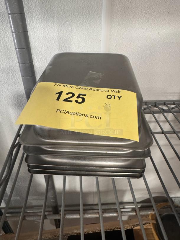 Stack of stainless steel food pans, model BIDX5, on a metal shelf. Quantity of five indicated on yellow label.