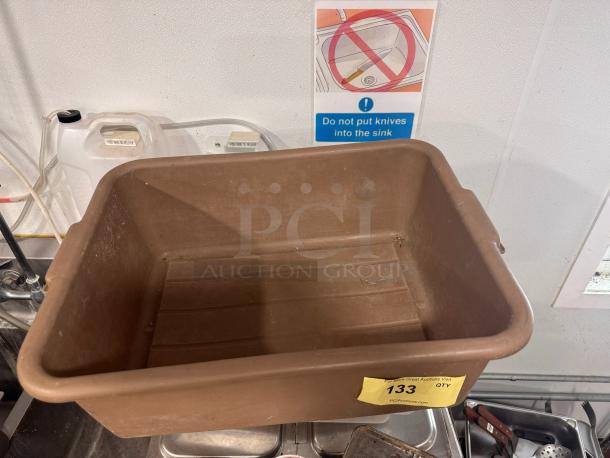 Brown plastic bus tub with handles, slight wear visible, auction label "133 QTY," and advisory sign in background.
