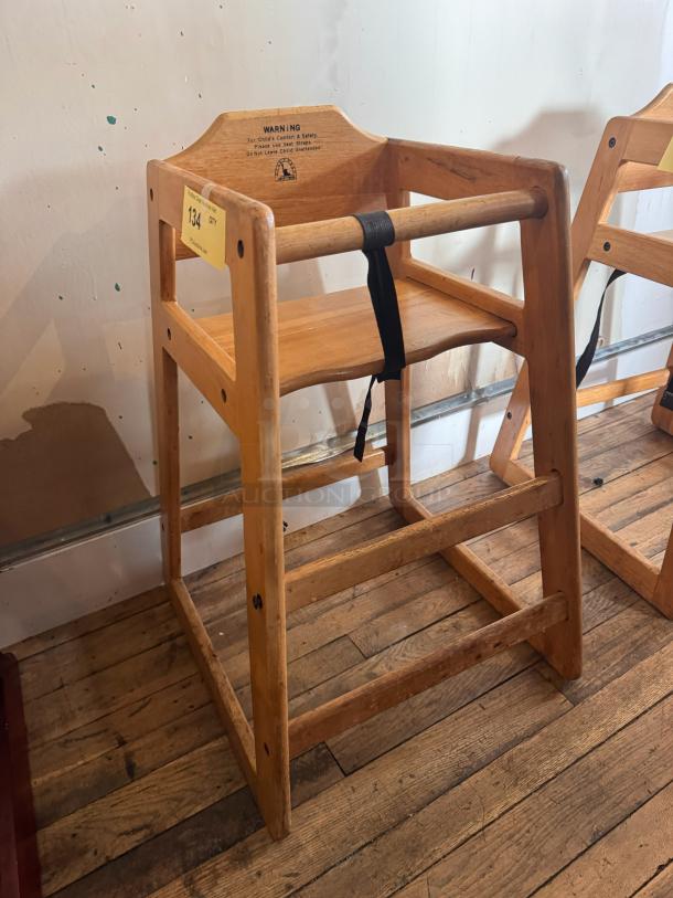 Wooden high chair with sturdy construction, safety strap, visible wear. Features a safety warning label on the backrest.