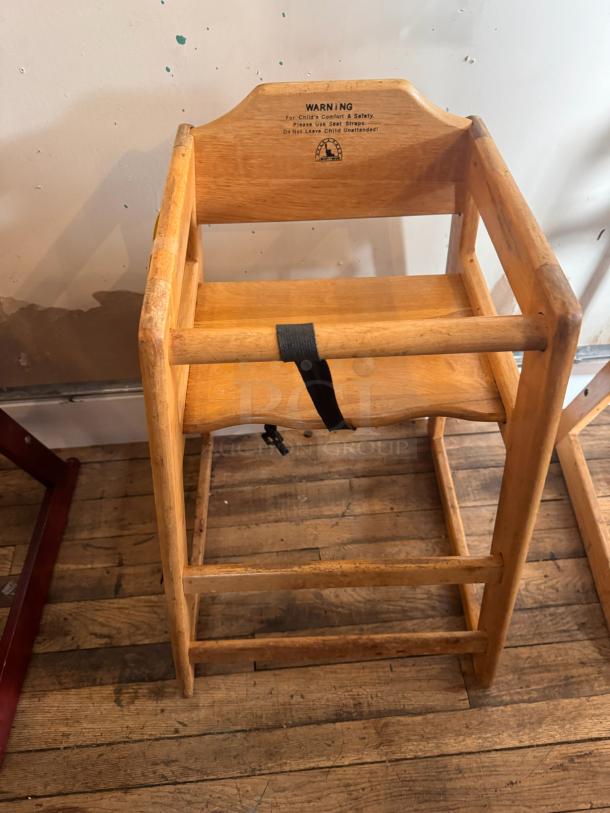 Wooden high chair with armrests and safety belt, shows wear. Warning label advises using seat straps for child safety.