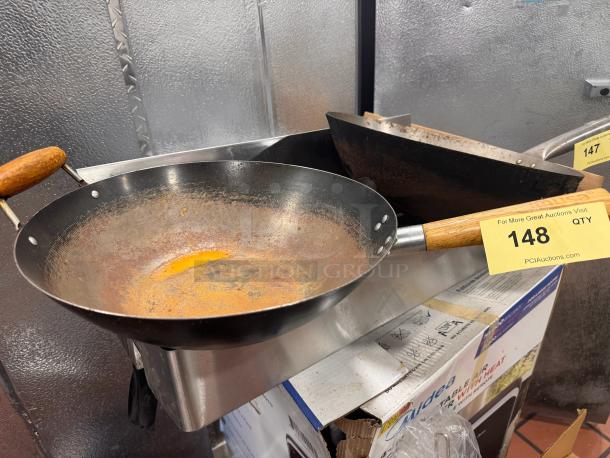 Rustic fry pan and wok with wooden handles, showing signs of wear and rust. Auction tag "148" from PCI Auctions visible.