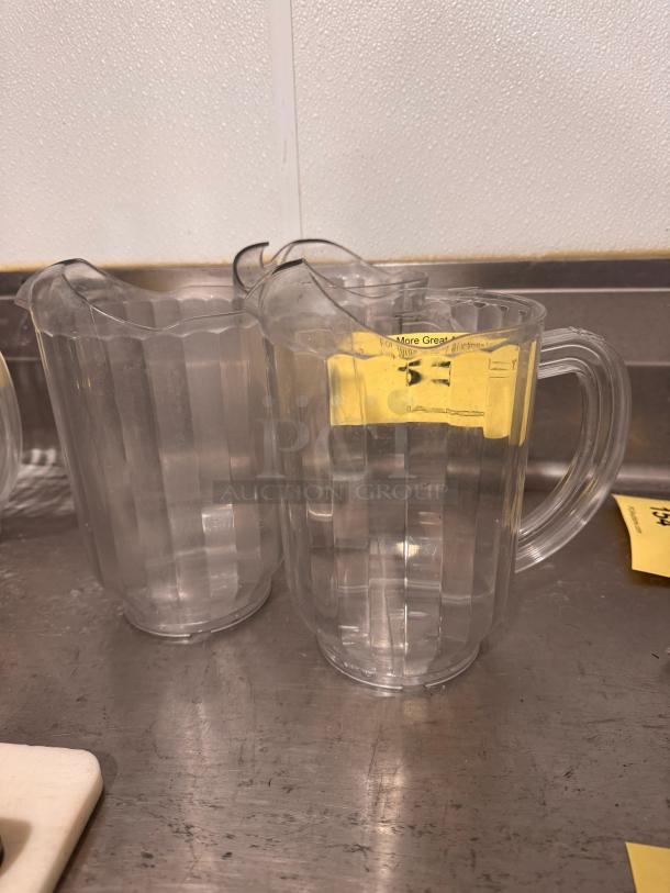 Two clear plastic pitchers, BIDX3, with ribbed design and integrated handles, in good condition, ideal for beverages.